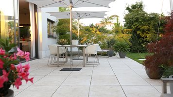  Terrasse mit weissen Bodenplatten, weißen Gartenmöbeln und zwei Sonnenschirmen. Abendsonne.