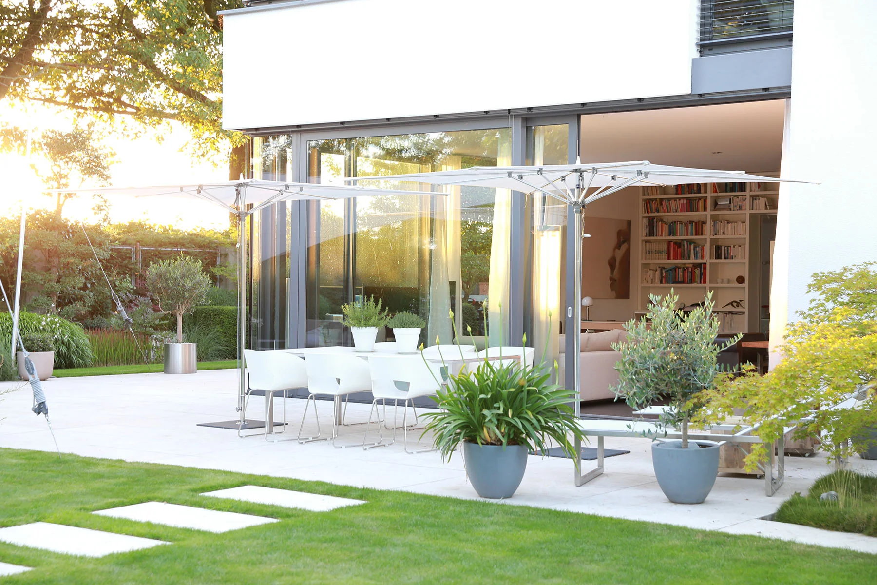  Terrass mit Gartenmöbeln, Blumenkübeln, Bodenplatten und Sonnenschirm. Die Sonne scheint.