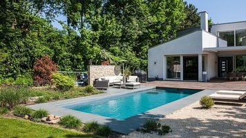 Ein Swimming Pool, eine Terrasse aus grauen Schiefernplatten, einem Beet aus Kies, zwei Liegestühle und einem Haus.