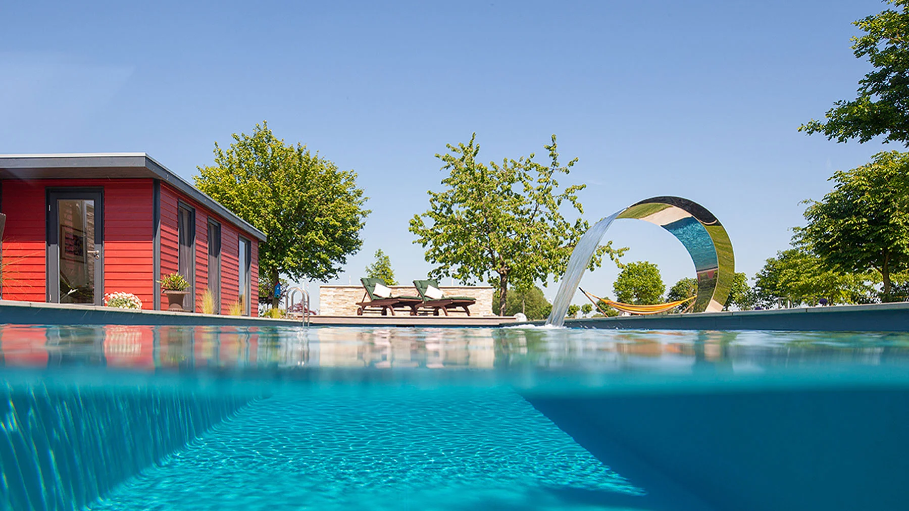 Ein großer Living Pool, eine Sauna aus roten Holzplatten, ein großer, grauer Sonnenschirm, Büsche und Pflanzen an der rechten Seite.