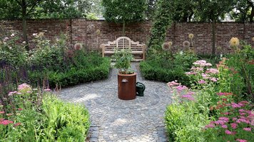 Garten für genießer. Ein gepflasterter Platz umgeben von Beeten. In der Mitte steht ein Kübel und eine Gießkanne.
