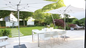 Terrasse für Designerfreunde. Eine weiße Terrasse, Gartenmöbel und zwei Sonnenschirme. Die Sonne scheint.